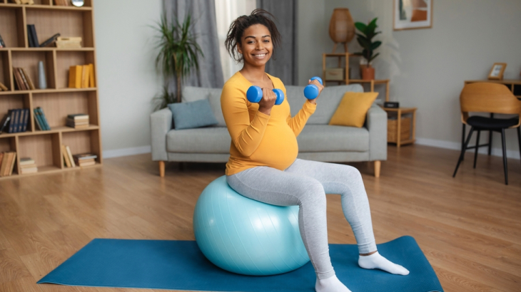 Mulher sentada na bola de pilates segurando 2 pesos